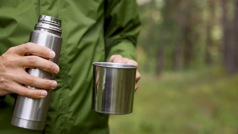 man pours hot tea from thermos flask into a mug in forest. nature adventure, tourism and camping concept