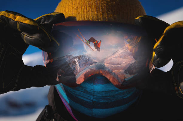 Close-up portrait of a skier in a mask holding on to ski goggles in the reflection of which the skier does a backflip trick against the backdrop of snowy epic high mountains stock photo