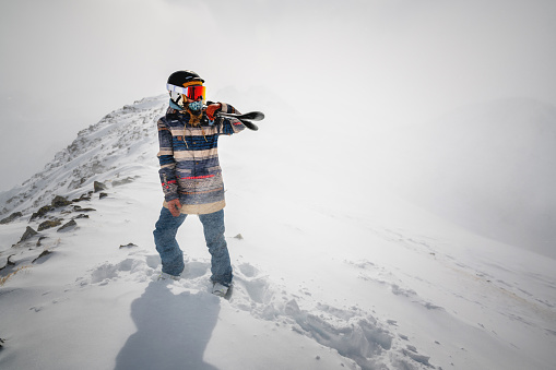 Full length shot of a cheerful skier carrying her skis on her shoulder. A skier is standing on top of a mountain and enjoying the view of the beautiful winter mountains on a cloudy day.