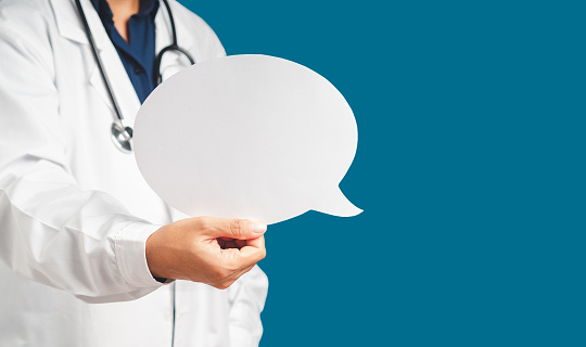 Midsection of doctor in uniform holding a white blank speech bubble while standing on a blue background. Space for text. Healthcare and advertising concept