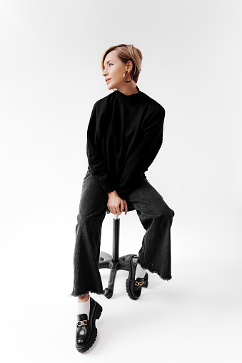 Full body of a woman dressed in a simple black outfit. She sits on a black stool against a white background, creating a contrast and emphasizing her minimalist style