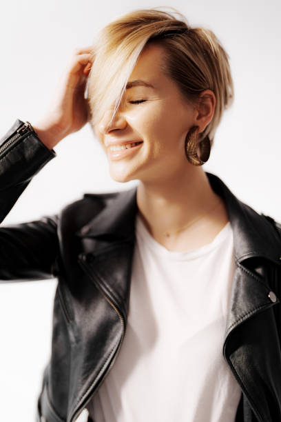 A happy blonde woman in a leather jacket on white background. The woman has her eyes closed, fair skin, and short straight hair. She looks shy and cheerful