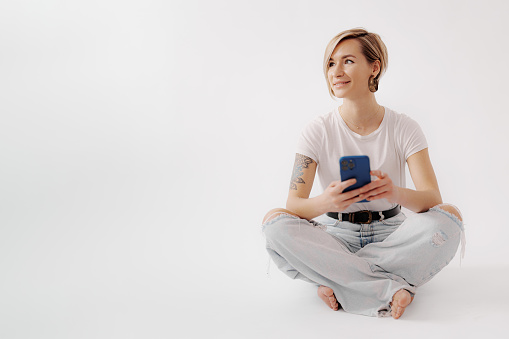 Young woman sitting on the floor holding a smartphone in her hands. She is wearing a white t-shirt and light blue jeans, and has a tattoo on her right arm. She looks relaxed and happy