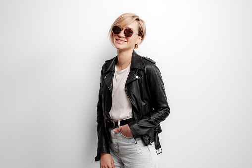 Fashionable young woman in a black leather jacket and sunglasses. She is standing against a white wall and smiling. She has a confident and stylish expression on her face