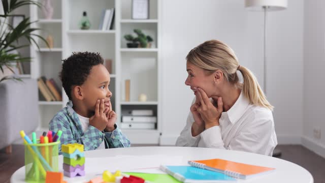 Cute little multiracial boy with speech problems practicing with occupational therapist, practicing and looking at mirror in office. The concept of childhood autism.