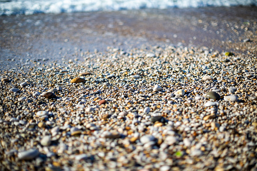 A pebble beach with wet and glistening pebbles of various sizes and colors, including white, gray, brown, and black. The background shows the ocean waves crashing onto the shore. The image is taken from a low angle, focusing on the pebbles in the foreground.