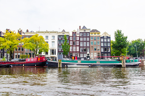 A canal in Amsterdam with boats and buildings in the background. The canal is lined with trees and the water is calm. There are several boats in the canal, including a red and a green one. The buildings in the background are tall and narrow, with different architectural styles and colors. The sky is overcast, and the overall mood of the image is peaceful.