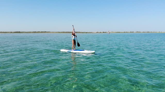 Aerial view from drone: young attractive woman in bikini and sunglasses floating on paddle board on turquoise water