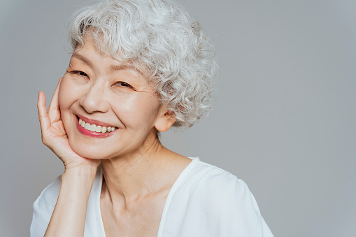 A senior Japanese woman is smiling and looking at the camera while touching her cheek.
