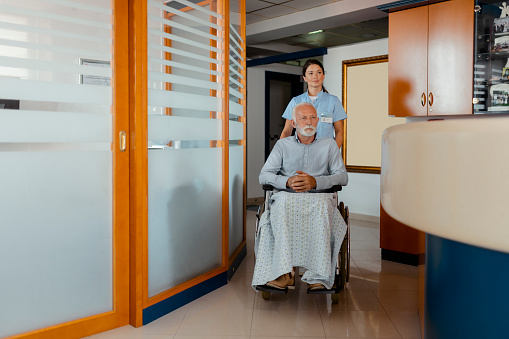 Female Caregiver Supporting Disabled Older Man Sitting in Wheelchair in Hospital