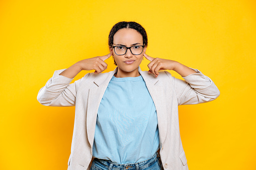 Displeased upset mixed race brunette business woman with glasses, company worker, covering ears with fingers, sadly looks at camera, stand on isolated yellow background