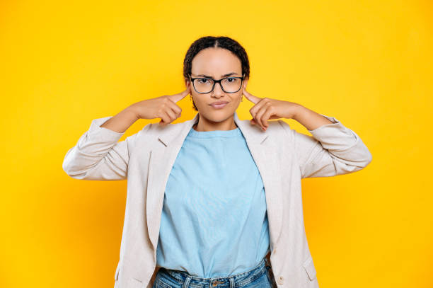 displeased upset mixed race brunette business woman with glasses, company worker, covering ears with fingers, sadly looks at camera, stand on isolated yellow background - silence finger on lips businesswoman one person imagens e fotografias de stock