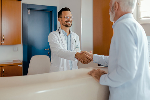 Young Doctor Shaking Hands With Senior Patient at Hospital Reception Desk. Medicine, People and Healthcare Concept