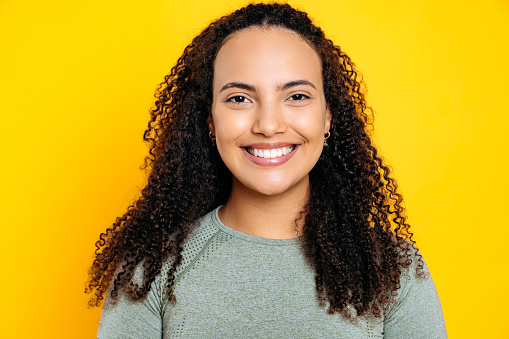 Close-up photo of beautiful positive smiling young mixed race woman with curly hair, dressed in a sporty t-shirt, standing on isolated yellow background, looking at camera, with toothy smile