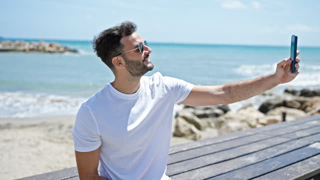 Young hispanic man smiling confident making selfie by the smartphone at seaside