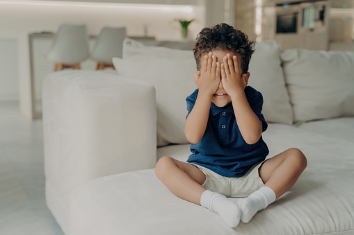 Excited boy in t-shirt and shorts plays hide and seek, covering his face, eagerly waiting to start searching, on a white couch in a modern room.