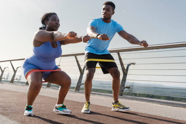 African american body positive woman in sportswear exercising outdoors, squatting with fitness band Attractive african american body positive woman in sportswear exercising outdoors, squatting with fitness band. Young trainer training plus size female, helping. Body transformation concept body positive couple stock pictures, royalty-free photos & images