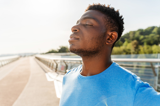 Portrait of attractive Nigerian man in blue t shirt standing on city street with closed eyes, relaxing after training. Handsome young guy posing for picture with copy space