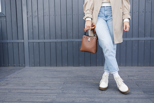 Fashionable young woman closeup in a trendy beige coat in blue jeans, white boots with a brown handbag walking down the street of the city. Autumn lifestyle style for a sunny autumn day. No face