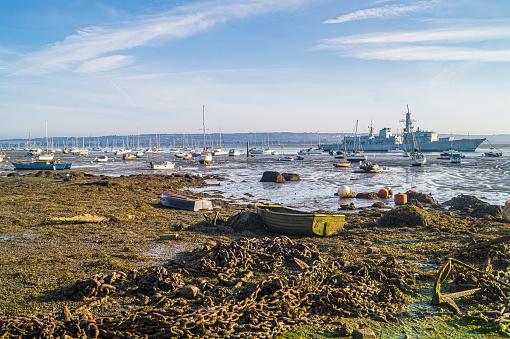 A view of Portsmouth Harbour taken from Gosport, looking east. Various shipping can be seen including yachts, boats and naval vessels. A decommissioned Royal Navy Frigate forms part of the scene.  In the foreground old chains and anchors litter the seabed, exposed at low tide.