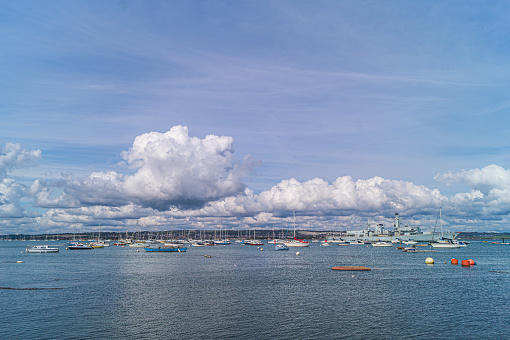 A view of Portsmouth Harbour taken from Gosport, looking east. Various shipping can be seen including yachts, boats and naval vessels. A decommissioned Royal Navy Frigate forms part of the scene.