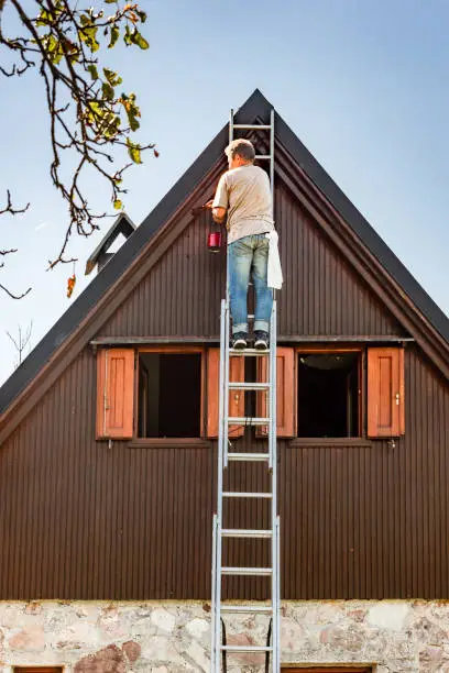 Photo of Man on Ladder Protecting Wooden House Exterior  Applying Paint with Paintbrush