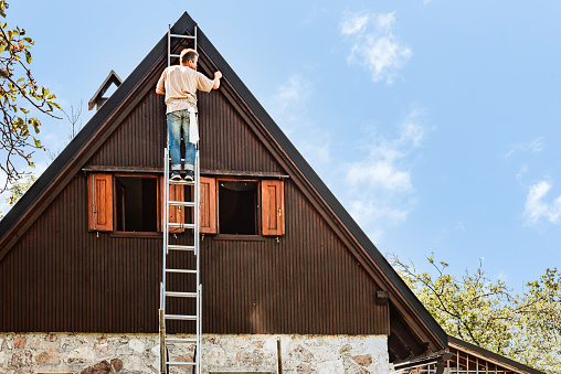 Man on Ladder Protecting Wooden House Exterior  Applying Paint with Paintbrush