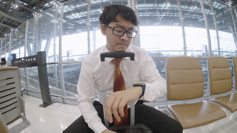 Businessman putting his chin on luggage handle, checking the time on his watch using smart phone while worriedly waiting for his colleague at Suvarnabhumi airport in Bangkok.