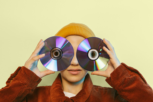Girl with yellow cap holding two compact discs over her eyes with colored background