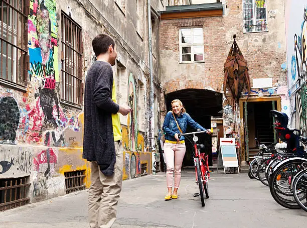 A young adult Caucasian woman is walking with her bicycle towards her boyfriend in a grafitti covered colorful alley.
