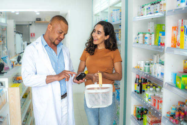 hombre farmacéutico afroamericano con cabeza rapada y barba, explicando la indicación del medicamento a una hermosa paciente latina en la tienda farmacéutica moderna. farmacéutico afroamericano profesional que recomienda al cliente para la dosificació - pharmacy pharmacist customer latin american and hispanic ethnicity fotografías e imágenes de stock