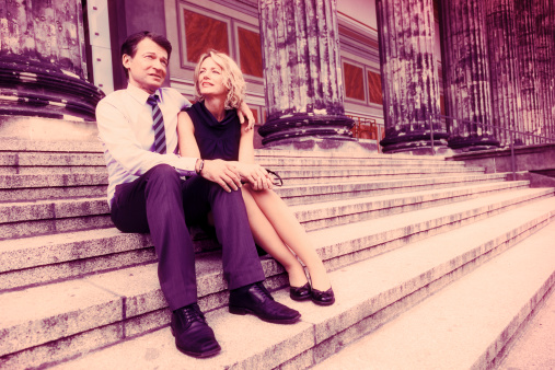 Elegant Caucasian mature couple in their early 50's sitting together on stone steps at a classic looking building facade. Image stylized with 'cross-processed' (Xpro) aesthetics.