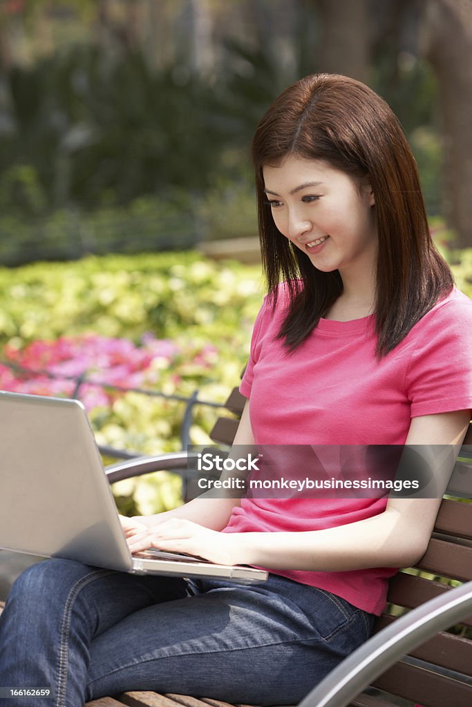 Chino mujer joven usando computadora portátil mientras se relaja en banco del parque - Foto de stock de 20 a 29 años libre de derechos