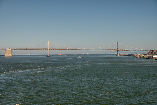 San Francisco, CA, USA - July 13, 2023: SF-Oakland Bay bridge entire western part with midway base and 2 towers. Seafaring ships on horizon under light blue sky. Trucks on top