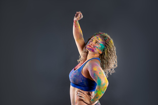 Bodypainting, creative makeup, bright colorful body art on gray studio background, plus size fat woman painted with powder paints