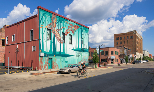 Eau Claire, Wisconsin, USA - August 3, 2023: Bicycle rider in front of colorful mural on turquoise wall at the intersection of Gibson St and Graham Avenue