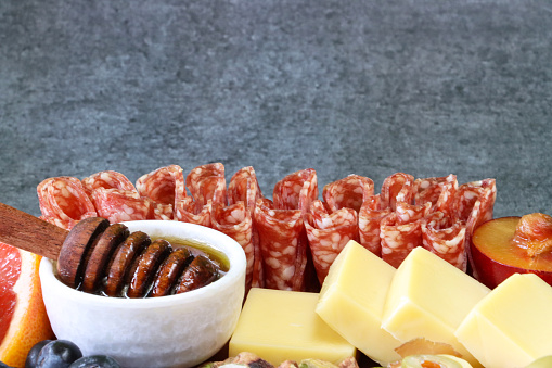 Stock photo showing close-up view of Halloween design charcuterie and cheese board covered with prepared sliced and chopped ingredients including folded salami slices, wooden honey dipper and ramekin of honey, plums, blueberries, pink grapefruit slices and cheddar cheese cubes.