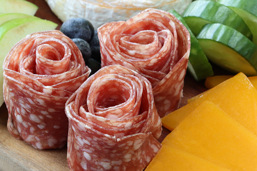Stock photo showing close-up, elevated view of Halloween design charcuterie and cheese board covered with prepared sliced and chopped ingredients including  salami roses, cheese slices, brie, cucumber, blueberries and green apple slices.