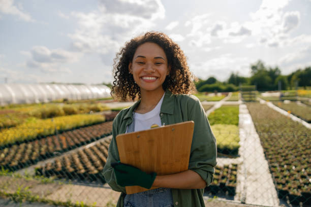 Positive woman farmer holding clipboard while standing on garden center background stock photo
