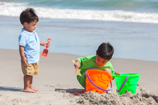 Two happy young hispanic boys brothers playing together on a sunny tropical beach with buckets and spades making sandcastles