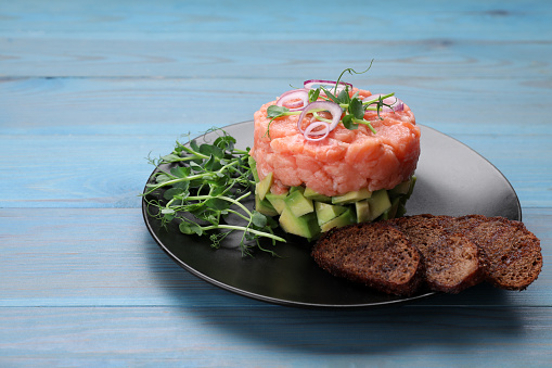 Tasty salmon tartare with avocado, microgreens and croutons on light blue wooden table. Space for text