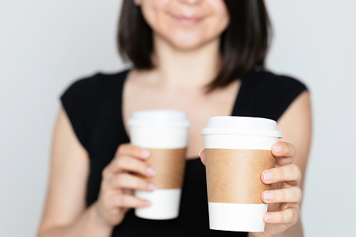 Unrecognizable caucasian businesswoman is  holding two disposable cups. One is for herself in the other cup is for a person in front of her.  She shows one of the cups to camera.