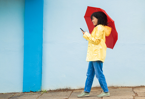 Woman, raincoat and phone typing with umbrella on city street for social media, internet or urban communication. Smile, happy and walking student with mobile technology for weather app check for rain