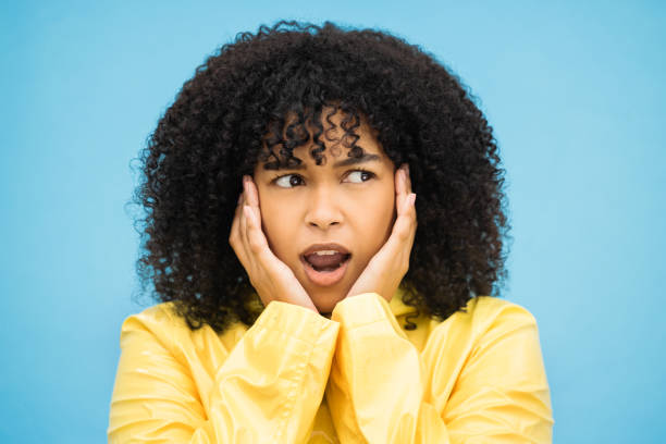 black woman, afro or shocked face on isolated blue background in raincoat weather change, rainfall or rain. model, person or surprised student with hands on cheeks in wow, emoji or scared expression - awe black ceremonial makeup stage makeup imagens e fotografias de stock