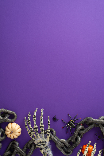 Spine-chilling Halloween scene: Vertical top view shot of thematic setup, captive's chain, eerie insects including spiders, zombie skeleton hands, pumpkins on violet background, space for greeting