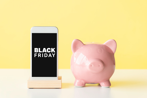 Front view of a smart phone and pink piggy bank on white table in front of a yellow wall. Black Friday text on device screen.