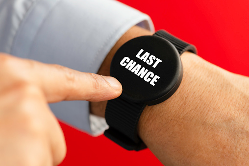 Businessman is using smart watch in front of a red wall. Last Chance  is written on the watch with white font.