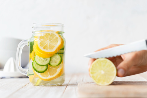 Infused water bottle with an abundance of lemon and cucumber slices on white wooden table. Hand of an unrecognizable person in the background is about to cut more lemon slices.