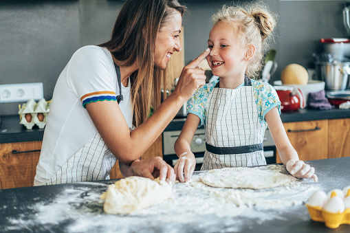 Cute little girl and her beautiful mom in aprons are playing and laughing while kneading the dough in the kitchen. Little girl kneading cookie dough in kitchen with mum. Happy mother and little girl with messy dough hands making biscuits on kitchen counter