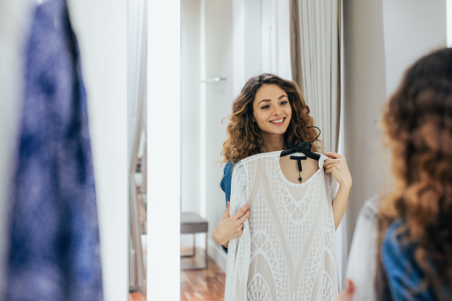 Young woman trying on clothes in a dressing room. Female client choosing dresses in luxury boutique. Beautiful woman get dressed up in the fitting room and looking at the mirror.